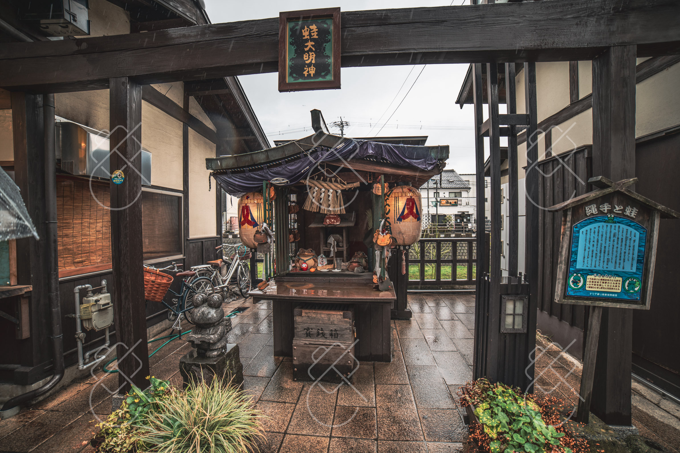 A roadside shrine sheltered under a wooden frame on a wet Matsumoto street — Matsumoto, fine art travel photography print available from Clever Tourists